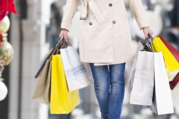 A shopper in a beige trench coat carrying several white, yellow and red paper shopping bags