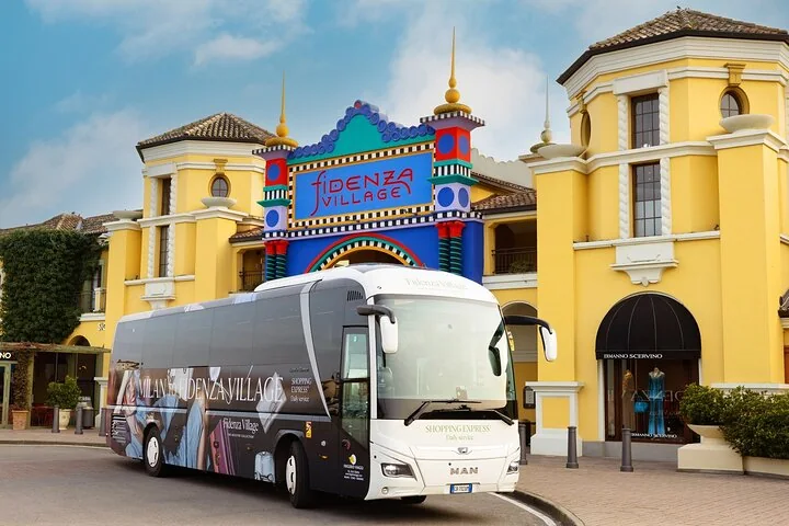 Un pullman bianco e nero parcheggiato davanti all'arco d'ingresso in piastrelle colorate di Fidenza Village tra edifici gialli