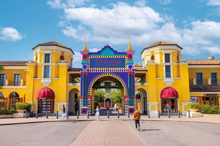 The colourful tiled entrance arch of Fidenza Village between two yellow towers under a blue sky